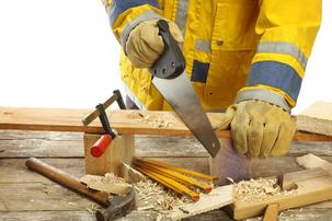 This is a picture of a carpenter with gloves and a yellow reflective jacket on sawing a piece of wood.